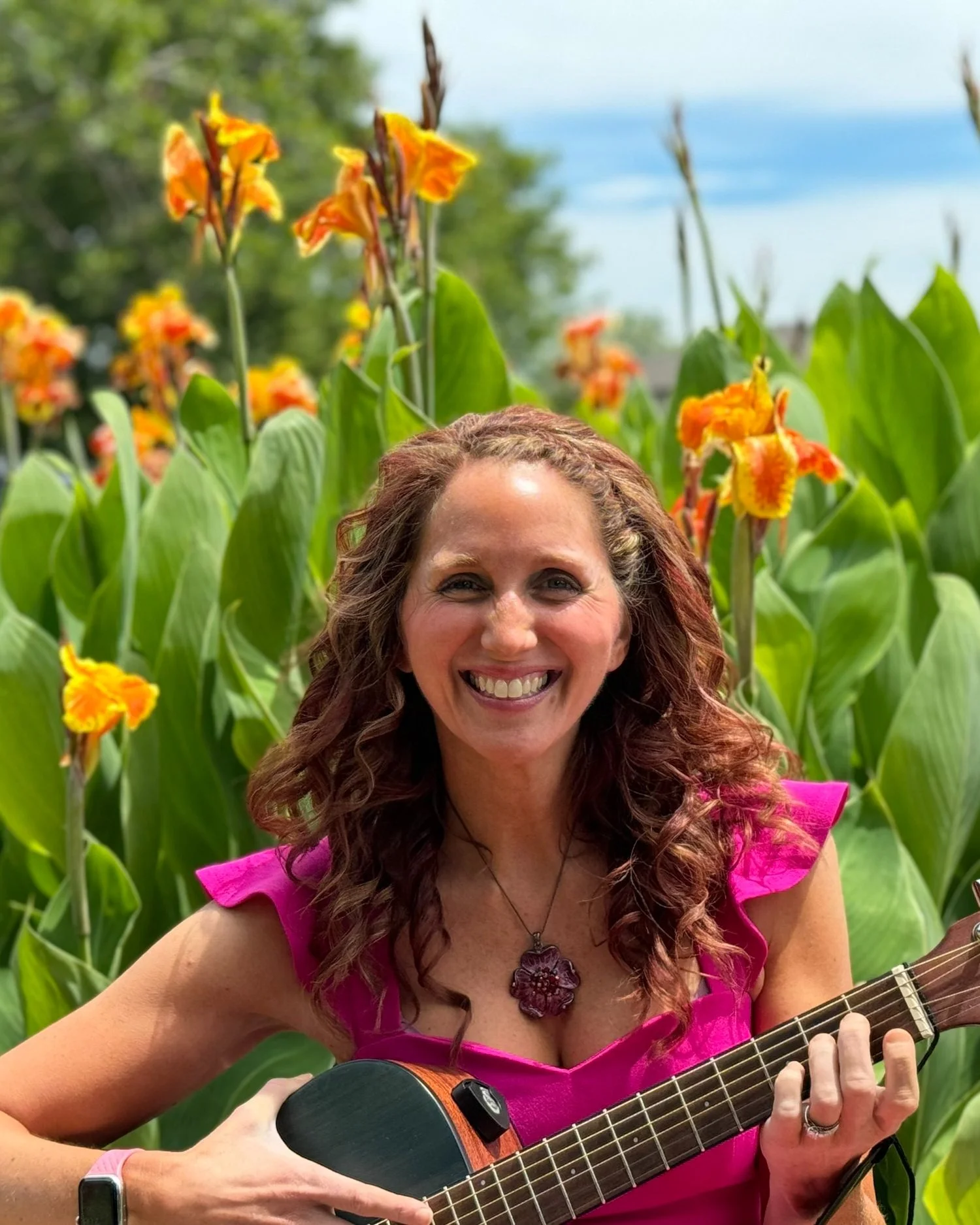 A woman with curly auburn hair smiling and playing guitar in a garden of tall orange flowers and green foliage.