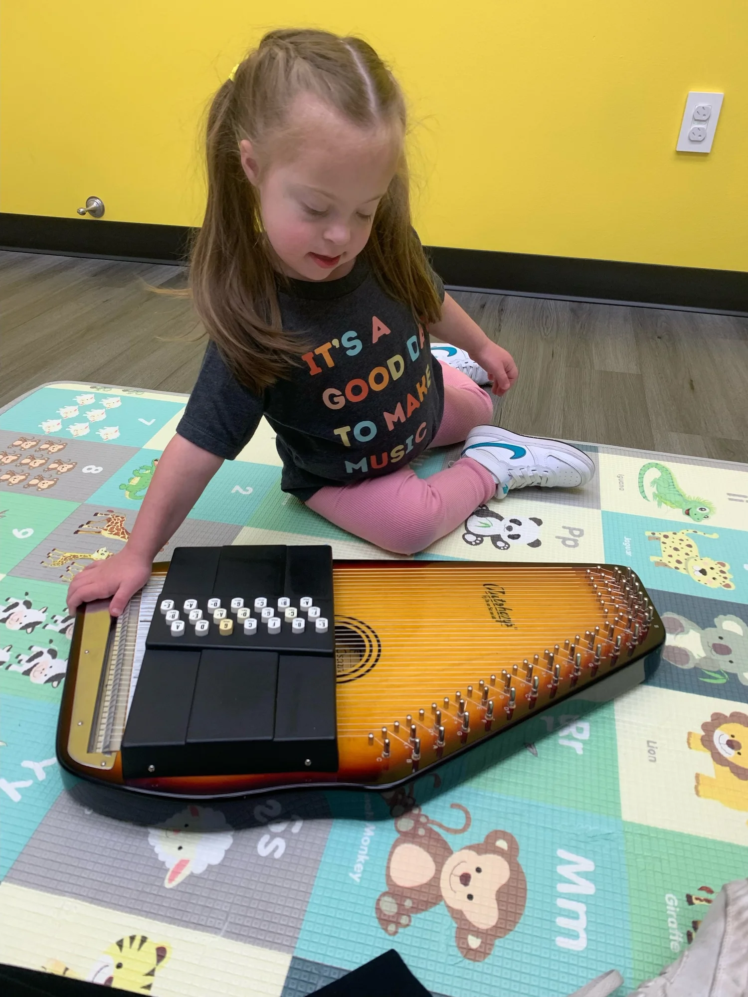 A young girl with brown hair, wearing a dark T-shirt and pink pants, sitting on a colorful animal-themed floor mat, playing an autoharp.