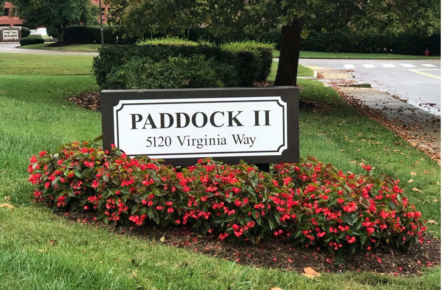 A sign reads "PADDOCK II, 5120 Virginia Way" next to a flower bed with red flowers, on a grassy area near a sidewalk and street lined with trees.