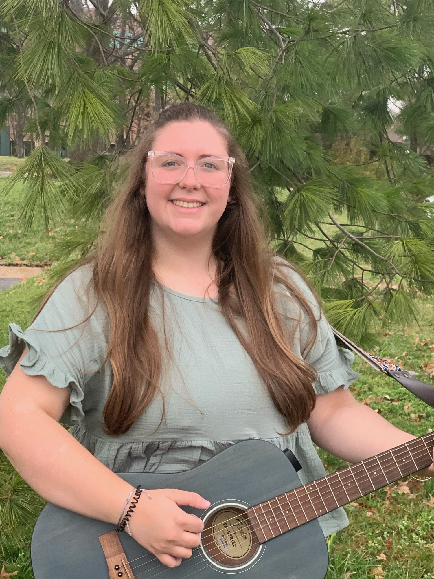A smiling young woman with long brown hair, wearing clear glasses and a light gray ruffled short-sleeve top, is outdoors playing a black acoustic guitar. She is sitting under a green pine tree, with trees and a grass lawn in the background.