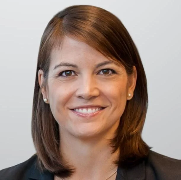 A woman with shoulder-length brown hair smiling at the camera, wearing a black blazer and pearl earrings, with a plain white background.