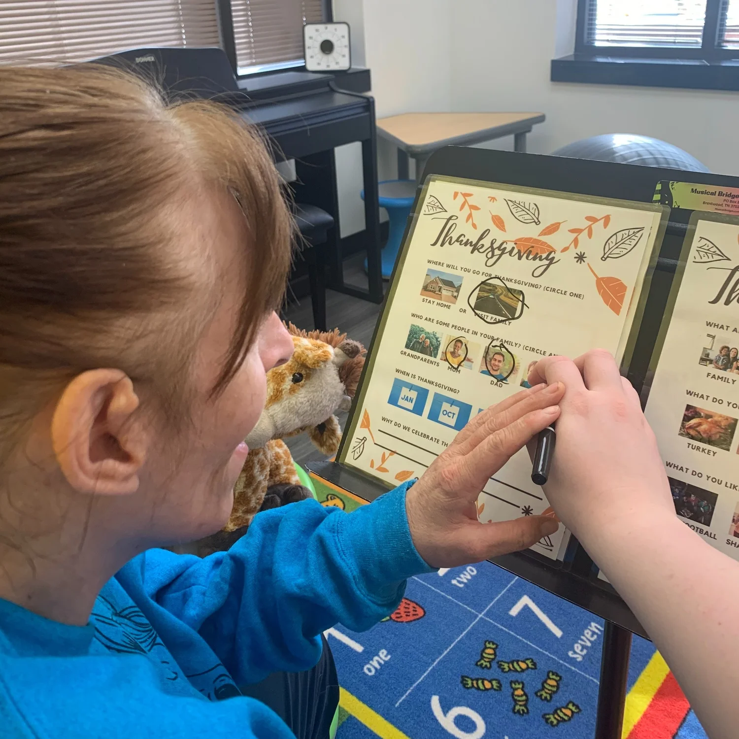 Young girl with red hair wearing a blue shirt, sitting and coloring in a Thanksgiving-themed worksheet at a table with a plush giraffe toy nearby.