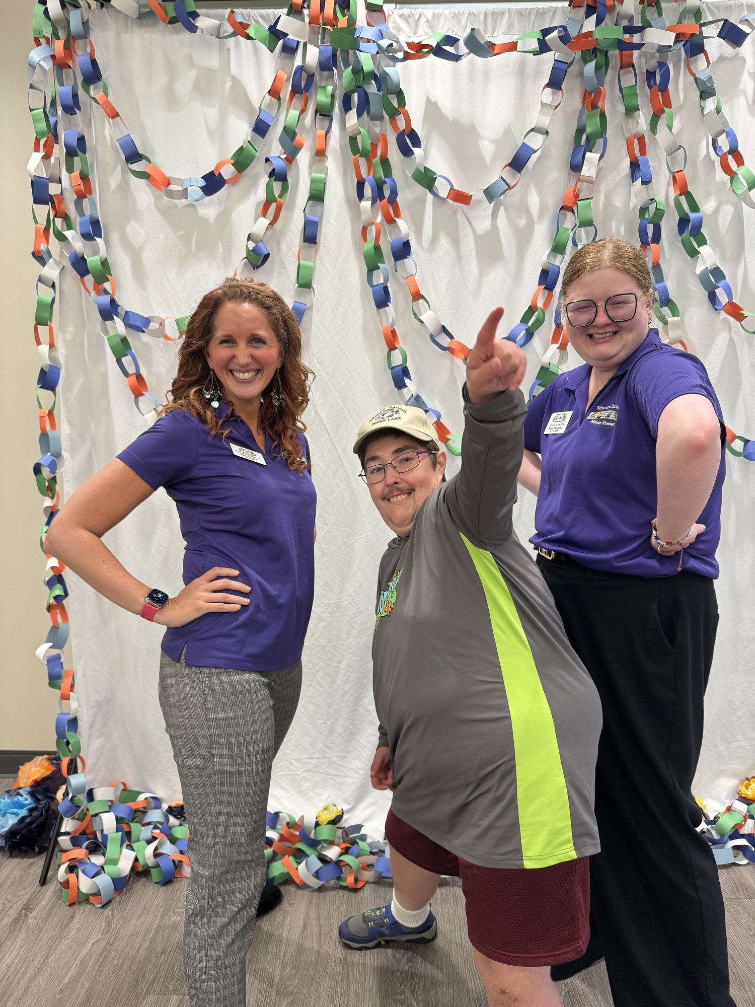 Three smiling people posing in front of a paper chain decoration backdrop, with one pointing upwards and the others smiling, at an indoor event.